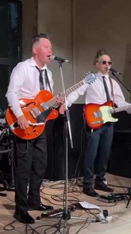 The video shows two male musicians performing on a stage. The musician on the left is singing into a microphone and playing an orange electric guitar. He is wearing a white dress shirt and a black tie, with black pants and black shoes. The musician on the right is also singing into a microphone and playing a red and orange electric guitar. He is wearing a white dress shirt and a black tie, with dark pants and dark shoes. He is bald and wearing sunglasses. The stage appears to be indoors, with a neutral-colored wall behind the musicians. There are microphones, guitar cables, and music equipment on the floor in front of them. The lighting is dim, suggesting an evening performance.