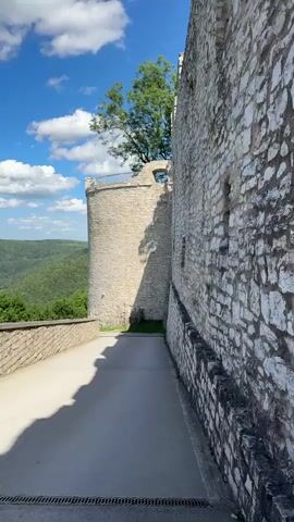This video shows a paved pathway leading around the exterior of a stone castle. A round tower is visible on the left, and a section of the castle wall with small windows runs along the right. Lush green hills and trees form the backdrop under a bright blue sky with white clouds. Sunlight casts shadows on the pathway.