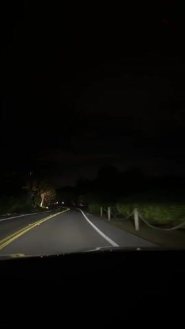 The video shows a car driving on a two-lane road at night. The headlights illuminate the road ahead, which curves to the left. There are trees and vegetation on either side of the road, and some lights can be seen in the distance. A house is visible on the right side of the road. The sky is dark and there are no stars visible.