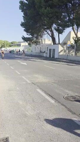 A group of cyclists are riding down a paved road on a sunny day. The road has white dashed lines marking lanes. On the left side of the road is a sidewalk with a person standing on it. On the right side of the road are buildings and trees. The cyclists are wearing helmets and athletic clothing. Some of the cyclists are close together, while others are spread out. The cyclists are moving from left to right in the frame. The camera appears to be mounted on a vehicle or held by someone walking alongside the road.
