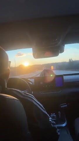 The video captures a serene moment inside a car, looking forward through the windshield. The back of the driver, wearing a dark jacket with white stripes on the sleeves, is visible in the foreground. The driver appears to be wearing glasses.

The car is traveling on a multi-lane highway during sunset. A brilliant, golden sun dominates the horizon directly ahead, casting a warm glow and producing lens flares on the windshield. The sky is mostly clear with a few clouds. Other vehicles, including large trucks, can be seen on the highway in the distance. Trees line the roadside, suggesting a natural landscape beyond the road.

Inside the car, a modern dashboard and steering wheel are visible. The steering wheel's digital display shows "110" and "107," likely indicating speed or distance. A prominent infotainment screen on the center console displays various icons, including what appears to be music playback controls (play/pause, volume), although specific text is unreadable. The interior of the car is dark, creating a contrast with the bright external light. The scene conveys a sense of peaceful travel during the golden hour in Tiush, Russia.