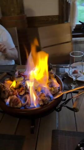 A dish with meat, onions, peppers, and other items is set on fire on a wooden table in a restaurant. The flames are tall and bright, with both orange and blue hues. A glass, plate, and cutlery are visible on the table next to the dish. Wooden chairs are partially visible in the background, with a window showing greenery outside. A portion of a person's arm can be seen in the upper left corner of the frame.