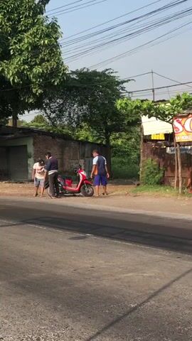 The video begins with a wide shot of a street scene. A red scooter is parked on the side of the road. Three men are standing near the scooter. The man on the left is wearing a white t-shirt and shorts. The man in the middle is wearing a blue jacket and dark pants. The man on the right is wearing a blue t-shirt and shorts. The man in the middle appears to be talking to the man on the left, and the man on the right is standing and looking towards the scooter. In the background, there are buildings, trees, and power lines. The lighting suggests it is daytime, possibly late afternoon due to the angle of the light.