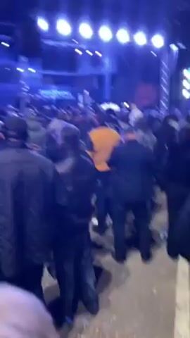 Here's a description of the video:

The video shows a large crowd of people gathered in an outdoor area at night. A stage is set up with bright blue lighting illuminating the scene. A large screen displays text and images, including a portrait of a man. The crowd is generally facing the stage, and some people are taking pictures or recording the event with their phones. The atmosphere appears to be somber and respectful. The event seems to be a memorial or commemoration, as indicated by the text on the screen.