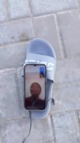 A phone lies on a gray sandal, connected to wired headphones. A video call is in progress on the phone screen, featuring a person wearing a brown beanie and a plaid shirt. The phone and sandal rest on a surface of gray paving stones. The video is shot from a top-down perspective.