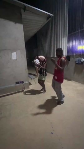 Three people are dancing outside a building under a metal awning. The person closest to the camera wears a red basketball jersey and grey pants. To his left, another person wears a black jersey and khaki shorts with a towel over their head. Behind them, another person in a white shirt and shorts also dances. The building has corrugated metal walls and a concrete floor. There's a bench near the wall to the left. It is dimly lit. The camera stays still for the duration of the clip.