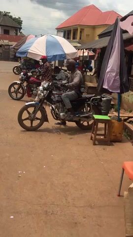 The video shows a daytime scene in Enyibichiri, Nigeria, on an unpaved, reddish-brown ground, resembling a street or market area.

Two men are prominently featured, each seated on a motorcycle. The man in the foreground is on a black motorcycle, wearing a grey long-sleeved shirt, blue jeans, and a grey cap. A black shoulder bag with "DIOR" text is visible across his chest. The other man is on a red motorcycle, dressed in a colorful patterned short-sleeved shirt, light-colored trousers, and a red baseball cap. Both motorcycles have "TOP TA" visible on their fuel tanks and feature multi-spoke wheels.

The setting includes two large umbrellas providing shade: one with blue and white stripes and another black and pink/purple. A small green wooden stool and a yellow jerry can are positioned near the black umbrella. In the background, there are buildings with red roofs, suggesting a commercial or residential area. Other motorcycles and people are visible further down the street, indicating an active environment. The weather appears sunny, and the overall mood is casual, with the men seemingly waiting or observing their surroundings.