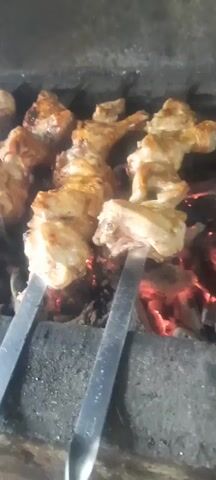 The video shows chicken pieces on skewers being grilled over hot coals. The chicken is browned and appears to be cooking. Smoke is visible rising from the grill. The skewers are made of metal. The background is dark and indistinct.