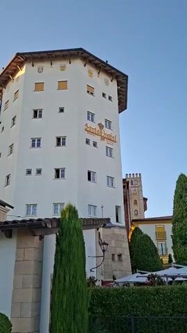 The video shows a tall, octagonal white building with a dark roof. The building has many windows and a sign that says "Santa Isabel" in gold lettering. There are several flags or crests above the windows on the top level. A smaller, connected building is visible to the left, also white with balconies and windows. In the background, another tower-like structure with a crenelated top is visible. A tall, green, conical tree stands in front of the white building, and a low wall with stone accents runs across the foreground. The sky is clear and blue.
