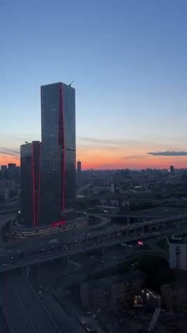 This is a vertical aerial drone video of a city skyline at sunset. The main subject is a very tall, modern skyscraper with red lights accenting its edges. The skyscraper is surrounded by a complex network of elevated highways with cars driving on them. In the distance, there is a dense urban landscape with many buildings and a vibrant orange and pink sunset sky. The camera is moving slowly upwards and to the right, providing a sweeping view of the cityscape and the setting sun. There are no people or specific actions visible, only the static urban environment and the dynamic sunset.