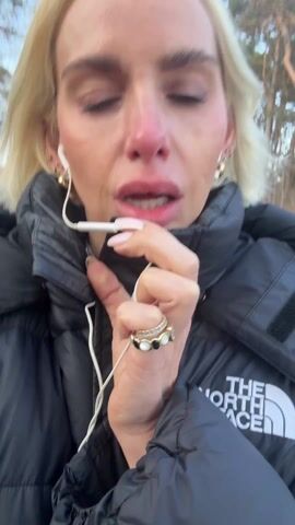 Here is a brief description of the video:
The video shows a woman with short blonde hair wearing a black North Face puffer jacket. She's outside, possibly in a park or wooded area, and holding her earphones up to her mouth as she speaks. She has earrings and several rings on her fingers. The woman looks directly at the camera at several points.