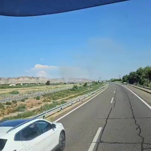 The video shows a view from a vehicle traveling on a highway. The highway curves to the right and is bordered by a metal guardrail and vegetation. In the distance, there are hills and a clear blue sky with some clouds. A white car is traveling on the highway in the same direction as the camera. There is a plume of smoke visible in the distance, rising from behind some structures. The smoke appears to be coming from the area of the hills. There are also some other vehicles visible on the highway and some electrical poles. The overall scene suggests a dry, possibly rural or semi-urban environment under a bright sky.