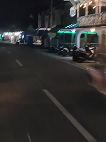 The video shows a nighttime scene on a road. A person is riding a motorcycle, facing away from the camera. The motorcycle has its brake light on. On the right side of the road, there are several motorcycles parked. On the left side of the road, there are stalls or shops lit up. There are other vehicles and people visible in the distance. The overall impression is of a busy street at night, possibly a market or a commercial area.