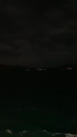 The video shows a dark, cloudy sky over a body of water. Distant lights from a town or city can be seen on the horizon. The water appears choppy, with some whitecaps visible in the foreground. The overall scene is dim and atmospheric, suggesting it was filmed at night or during twilight.