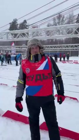 A man wearing a fur hat, a grey jacket, black pants, and a red vest with the word "СУДЬЯ" (judge) on it, is standing in the snow and facing the camera. He is surrounded by snow and a ski area in the background. There are people skiing and standing around in the distance, and a metal structure is visible overhead. The man is standing between red and white striped markers. The weather appears to be cold and overcast. The man pats his vest.