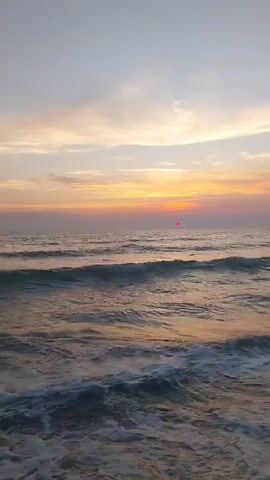 The video shows a sunset over the ocean. The sky is filled with soft clouds in shades of pink, orange, and yellow, with a pale blue gradient towards the top. The sun is visible as a small, red-orange orb just above the horizon. The ocean is calm, with gentle waves rolling towards the shore, creating white foam. The water reflects the colors of the sunset, giving it a warm, golden hue. The camera is positioned at a low angle, close to the water's surface, giving a sense of immersion in the scene. There are no people or man-made objects visible in the video. The overall atmosphere is peaceful and serene.