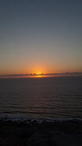 This is a vertical, wide-angle photograph of a sunset over the ocean. The sun is a vibrant orange orb, partially obscured by a thin line of clouds just above the horizon. The sky transitions from a deep blue at the top of the frame to a soft orange and pink hue closer to the horizon. The ocean is a dark, deep blue, with gentle waves lapping at the rocky shore in the foreground. The shore itself is dark and indistinct, providing a textured contrast to the smooth expanse of water and sky.

The image captures a serene and peaceful moment. The time of day is clearly sunset, indicated by the color of the sky and the position of the sun. The weather appears calm, with only slight ripples on the water. The location is coastal, with the vastness of the ocean meeting land. There are no people or man-made objects visible in the frame, emphasizing the natural beauty of the scene. The overall emotion conveyed is one of tranquility and awe at the natural spectacle. There is no visible text within the image.