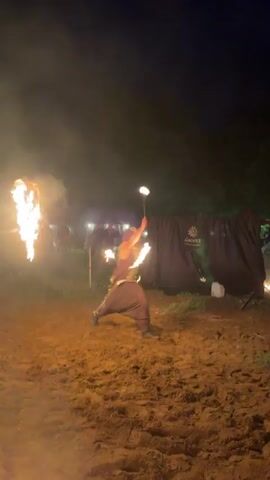 Two fire performers are seen at night on a dirt ground. A male performer in dark clothing with metallic accents breathes fire and manipulates various fire props, including spinning flames around his body. A female performer in a white dress and white boots manipulates a large fire fan with multiple flames. They move around the performance area, with distant tents and lights visible in the dark background.
