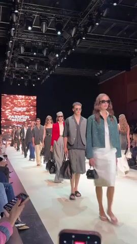 A fashion show is underway with models walking down a runway. A screen in the background displays the text "SUMMER FASHION SHOW". The models are diverse and wearing various stylish outfits, including suits, dresses, and separates, accessorized with sunglasses and handbags. The runway is white and well-lit, and the audience is seated on the left side of the frame, with some people holding up phones to record. The overall atmosphere appears to be professional and high-fashion.