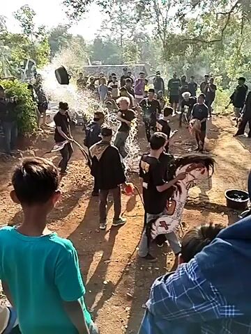 A crowd of people are gathered outdoors in a semi-rural setting. Some are standing and watching, while others are in the center of the gathering. There is dust or smoke in the air. Several people are wearing dark clothing, some with head coverings. One person appears to be holding what looks like a whip or rope. Another person is lying on the ground, and a few individuals are interacting with them or the objects around them. The environment appears to be a dirt path lined with trees and vegetation, with some rudimentary structures in the background. The time of day seems to be daytime, with sunlight casting shadows.