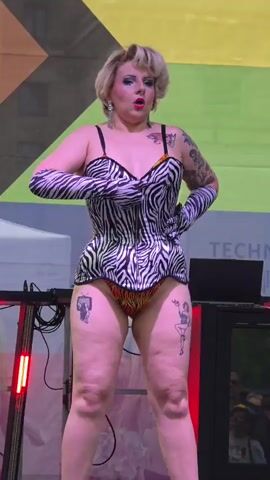 A blonde woman in a zebra-print corset and matching gloves is performing on a stage in front of a colorful backdrop. She has tattoos on her arms and legs. She is striking poses and interacting with the audience. There are DJ equipment and lights on the stage.