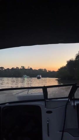The video shows a view from a boat at dusk, on a waterway filled with several other boats. The camera is positioned from inside the vessel, looking out through the windshield. The bow of the boat is visible in the foreground, with glimpses of the interior instrument panel. Through the windshield, several other boats are seen at anchor, mostly white in color, some with people visible. The sky shows a transition from light to dark, with a subtle orange glow on the horizon. The shore is lined with dense trees and vegetation. The boat appears to be moving slowly, gently rocking with the water. The lighting is soft, and the water reflects the colors of the sky. A dark canopy is visible over the camera. Loud music is audible in the background.