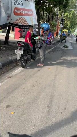 A young child in a red shirt sits on a black motorcycle parked on the side of a paved road. A person wearing a blue helmet, a striped backpack, and grey pants stands next to the motorcycle, holding a clear bottle, likely hand sanitizer. A large white banner with Indonesian text and imagery is visible behind the motorcycle. The environment appears to be an urban street with trees, traffic lights, and other vehicles in the background. The ground is asphalt with white lane markings.