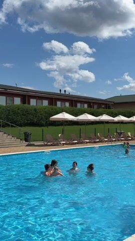 The video shows a bright, sunny day with a clear blue sky and fluffy white clouds. In the foreground, a group of children are playing in a swimming pool filled with clear blue water. They appear to be having fun and interacting with each other. In the background, there is a long, low building with a dark roof and many windows. A row of lounge chairs with closed umbrellas are set up on a green lawn in front of the building, suggesting a resort or hotel setting. The overall scene conveys a sense of summer recreation and enjoyment.