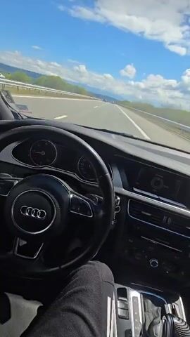 The video shows a first-person perspective from the driver's seat of an Audi car (identifiable by the logo on the steering wheel) traveling on a multi-lane highway. The driver's right hand briefly rests on the steering wheel at the beginning of the clip. A watch is visible on the driver's wrist, and their leg, dressed in dark trousers, is seen in the lower left.

The car's dashboard, instrument cluster, infotainment screen, and gear shifter are visible. The keys are hanging from the ignition.

Outside the car, the sky is bright blue with scattered white, fluffy clouds, indicating a clear, sunny day. The highway stretches ahead, appearing to be in good condition, flanked by safety barriers and green verges. In the distance, green hills or mountains line the horizon under the bright sky. The overall scene suggests a calm, pleasant drive. Music with a rhythmic beat and vocals is playing in the background. No specific text apart from the Audi logo is clearly legible. The scene is consistent with driving through a rural or semi-rural area, like the region around Dolna Dikanya, Bulgaria.