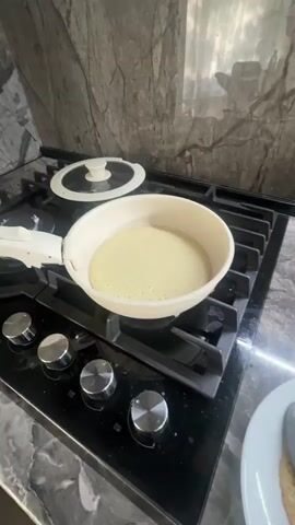 A person is cooking pancakes on a stovetop. The video begins with a close-up of a pan on a gas stove with pancake batter being cooked. Bubbles are forming on the surface of the pancake. The camera then pans to a plate with a cooked pancake and a spatula. Next, a bowl of pancake batter is shown, and the person uses a spoon to stir it. In the background, a kettle and a bottle of oil are visible. The video ends with a shot of the pancake batter in the bowl.