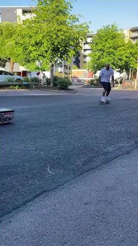A bald man wearing a white t-shirt and dark shorts is skateboarding in a paved area. He approaches a low, rectangular skate ramp with graffiti on its sides. He rides up the ramp and performs a trick, balancing on his skateboard atop the ramp. He then rides off the ramp and continues skateboarding on the paved surface. Cars and apartment buildings are visible in the background, along with trees. The sky is clear and blue.