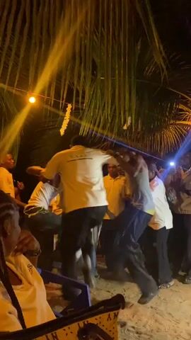 At night, under a canopy of palm fronds illuminated by warm yellow string lights, a lively group of young men is dancing outdoors. The ground beneath them appears sandy or dusty. Several men, mostly wearing light-colored shirts and dark trousers or shorts, are energetically moving together in a central circle, some with arms around each other's shoulders, showcasing various dance moves. Their expressions suggest enjoyment and camaraderie.

Around the main group of dancers, other individuals stand and watch, some swaying to the music. In the foreground, on the left, a person with braided hair is seated in a blue plastic chair, observing the scene. Another person is partially visible, also seated nearby. The overall atmosphere is festive and energetic, characteristic of a social gathering or celebration in Aného, Togo, with the tropical foliage adding to the specific locale's ambiance.