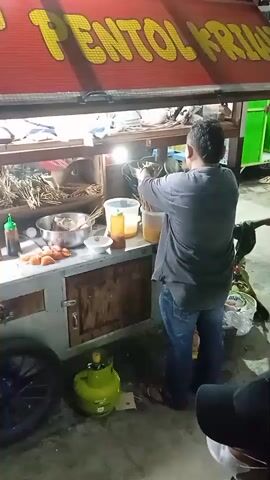 At night, an outdoor food stall is brightly lit by artificial lights. A man in a dark grey shirt and blue jeans stands behind the counter, actively cooking. He uses tongs to fry skewers of food in a pan or wok, which is heated by a flame from a green gas cylinder placed on the ground. The stall's red awning displays the yellow text "PENTOL KRI...". On the counter, there are various containers, including several with liquids, bowls, plates, bottles of sauce, and more skewers of food, likely pentol. Part of a motorcycle wheel is visible on the left, suggesting the stall is mobile or part of a motorbike. Another person, mostly obscured, is partially visible in the bottom right, wearing a black hat. The scene is set on a paved ground, typical of a street food vendor in Kebonsari, Indonesia.