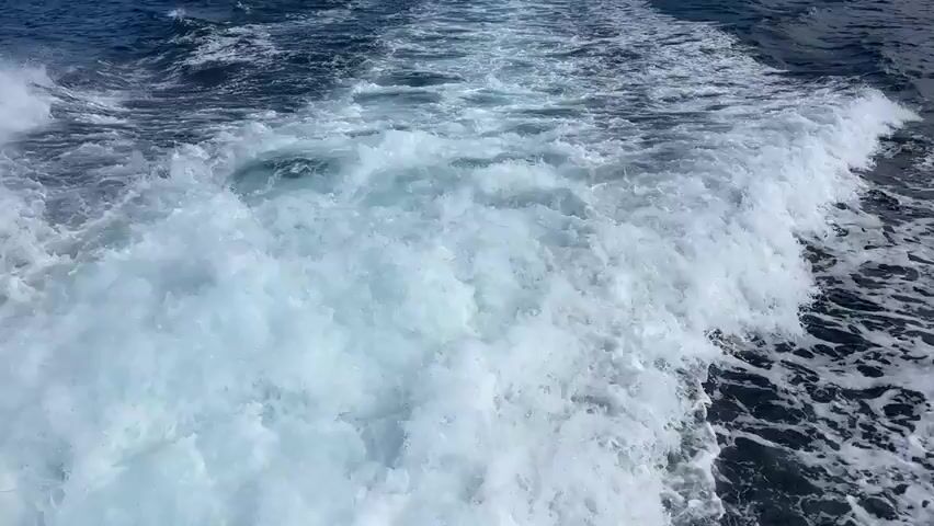 This video shows a wake behind a boat moving through dark blue water. White foam churns in the wake, creating a textured pattern. In the distance, a coastline is visible with green hills dotted with buildings. The sky above is blue with scattered white clouds. The camera appears to be positioned low, near the water level, looking backward from the moving vessel.