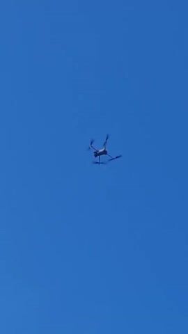 A small, dark-colored drone with four propellers is captured mid-flight against a clear, bright blue sky. The drone is positioned in the center of the frame, tilted slightly upwards and to the right. Its propellers are blurred, indicating movement. No other objects or people are visible in the frame. The lighting suggests it is daytime.