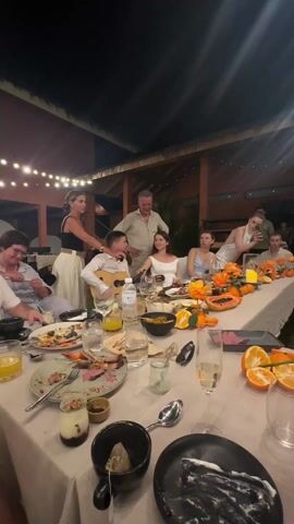 A well-lit outdoor evening scene shows a large group of people gathered around a long, messy dinner table with remnants of a meal: plates with leftover food, glasses, bottles of water, and discarded fruit peels. A young man sits at the table playing a guitar while a woman stands next to him, appearing to offer support or encouragement. An older man stands behind them, observing the performance. Other individuals at the table are engaged in conversation or looking on. The table is decorated with orange flowers and cut fruit, with string lights adding to the warm ambiance. Several people are standing in the background.