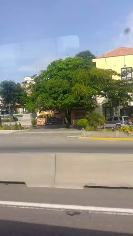 The video shows a view from a moving vehicle, likely a car, looking at a street lined with brightly colored buildings. The buildings are various heights and designs. Some have palm trees and other greenery in front of them. A concrete barrier separates the street from the road the vehicle is traveling on. The sky is blue and sunny. The Venezuelan flag is flying on a hill in the background.