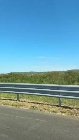 The video shows a view from a moving vehicle on a sunny day. The foreground features a gray guardrail on the right side of the road, with dry grass and some green foliage. The background consists of rolling hills covered in green vegetation and trees. The sky is a clear, bright blue with a few faint white clouds. The road surface is visible at the bottom of the frame. The camera appears to be mounted inside the vehicle, capturing the passing scenery.