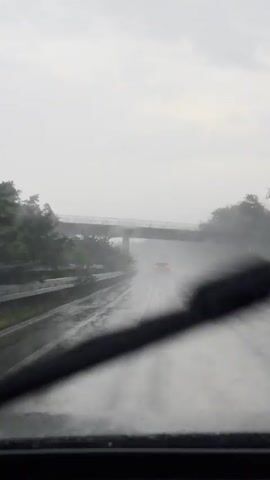 This video shows a view from inside a vehicle traveling on a highway during heavy rain. The windshield is wet, with raindrops visible. The highway is also wet and appears to have standing water in places, creating spray from the tires of other vehicles. A car ahead, with its brake lights illuminated, is driving in the same direction, producing a significant amount of spray. An overpass is visible in the distance, spanning across the highway. Trees and vegetation line the sides of the highway. The sky is overcast and gray.