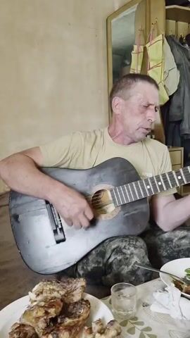 A man in a tan t-shirt and camouflage pants sits indoors playing an acoustic guitar. He is focused on his playing, with his eyes closed and a slight frown on his face. In front of him is a table laden with food, including plates of cooked meat, pickles, and what appears to be caviar. The setting appears to be a modest room with a neutral-colored wall and a closet filled with clothes in the background. The lighting suggests it is daytime. The man's demeanor suggests he is engrossed in his music, perhaps singing along. The presence of food implies a meal or a gathering, possibly a celebratory one given the spread.