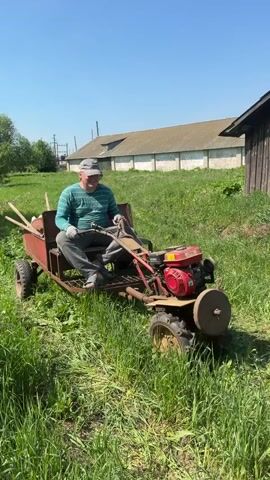 A man is driving a homemade vehicle across a grassy field on a sunny day. The vehicle consists of a small red engine, a metal frame with a seat, and four wheels. The man is wearing a green and blue striped shirt, gray pants, a gray hat, and blue sandals. He is smiling and looking at the camera. Behind him is a small wooden shed. In the background is a larger barn-like structure and some power lines. The grass is tall and green.