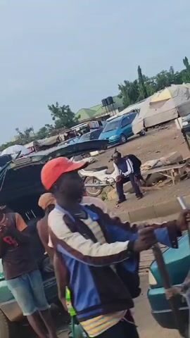 The video shows a crowded outdoor market scene in an urban area. Several people are visible, some walking, some standing around, and one pushing a wheelbarrow. Cars and motorized rickshaws ("keke napep" or "tuk-tuk") are parked around the periphery of the market area. There are many small stalls or tables set up, selling various goods, including fruits and drinks. The market appears to be located in a dusty area with a building visible in the background. There is a gas station, and various cars and a "keke napep" can be seen navigating through the area.