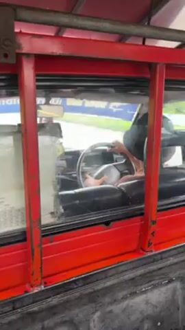The video shows the back of a red bus, the driver is visible through the window holding the steering wheel. The bus is moving along a wet road, other vehicles can be seen passing by. The back of the bus is open and has a metal frame with glass windows. A fare chart is visible on the left side of the window. The weather appears to be overcast.