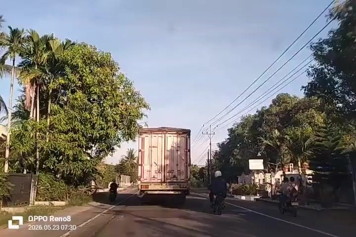 The video shows a view from inside a vehicle moving on a road. Ahead, a large truck with a boxy rear is traveling in the same direction. Two motorcyclists are also traveling ahead of the camera vehicle, one directly behind the truck and another further to the right. The road is asphalt and lined with trees and some buildings. Power lines run overhead. The sky is clear and bright. The video timestamp indicates a date of May 23, 2026 and a time of 07:30. The device watermark at the bottom corner indicates that the video was recorded on an OPPO Reno8.