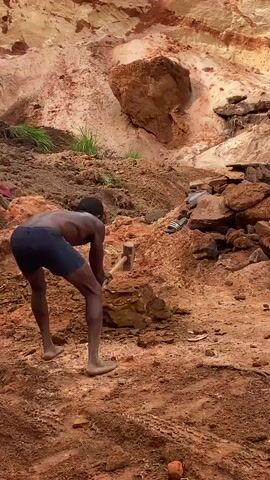 The video shows a shirtless dark-skinned man in shorts hammering at the ground in a quarry. The ground is red and the quarry walls are a mix of red, tan, and white. There are piles of rocks scattered around the ground. The sky is cloudy. The man is barefoot.