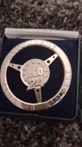The video shows a close-up shot of two medals inside presentation boxes. The first medal, on the left, is shaped like a steering wheel with the inscription "AUTOMOBILE CLUB DE L'OUEST" around the rim and "ACO 24" in the center. It appears to be made of a silver-colored metal. The second medal, on the right, is round and silver-colored with various embossed designs, including what looks like a wreath and some lettering that appears to be "UNC". The background of the video is a fuzzy, dark gray carpet. The lighting is dim, but the objects are clearly visible. The video is very brief, lasting only a few seconds, and the camera movement is minimal, primarily panning slightly to the right between the two medals.