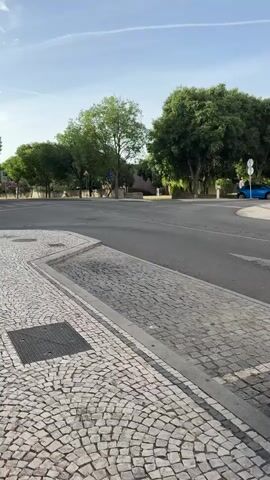 The video shows a view of a street intersection with traditional cobblestone sidewalks. In the foreground, the intricate pattern of the cobblestones is clearly visible, as is a metal grate embedded in the sidewalk. The street itself is paved and appears to be an asphalt road. On the far side of the intersection, a line of trees with lush green foliage creates a natural boundary. Beyond the trees, a building or buildings are partially visible. A bright blue car is parked or driving on the far side of the road, with its rear facing the camera. The sky above is a clear light blue, with a few faint white streaks that could be contrails from an aircraft. The lighting suggests it is daytime, likely morning or afternoon, with the sun casting shadows. No people are clearly visible or engaged in any significant actions. The scene is quiet and appears to capture a moment of stillness at a street corner.
