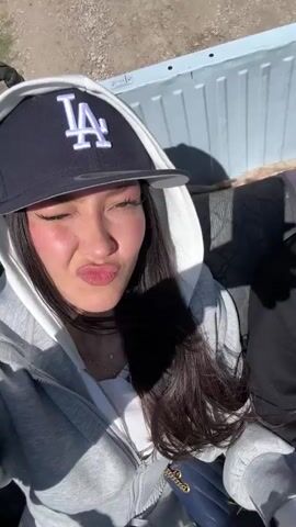 A young woman is the focus of this video. She is wearing a grey zip-up hoodie and a navy blue baseball cap with an "LA" logo. She is riding in the back of an open vehicle, possibly a truck or all-terrain vehicle, on a dirt road. Initially, she makes a pouting face. Then, she turns the camera to reveal a scenic view of green, tree-covered hills under a blue sky with fluffy white clouds. She smiles and opens her mouth wide in excitement. The top of the vehicle she's in is covered with black tape.