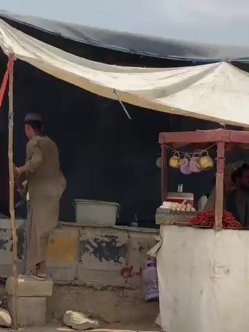 The video captures an outdoor food stall, likely situated in a market or roadside setting in Panjwai, Afghanistan, during daylight hours. The stall features a sturdy red-framed structure with a white lower facade and is covered by a white canvas canopy providing shade.

Several men are visible working at the stall. One man, wearing a dark shirt, stands prominently behind the counter, appearing to be the main vendor or operator. Another man in a lighter shirt or jacket is partially visible to his right, and a third individual in a white tunic is seen further to the right.

On the counter, various food items are displayed on plates. These include what appear to be bright orange, spiral-shaped sweets, characteristic of jalebi, and another plate holding yellow-colored items, possibly savories or different sweets. Small, possibly decorative, lights or ornaments hang from the stall's frame. The ground around the stall appears to be unpaved or dusty. The men are engaged in the activity of operating the food stall, presumably serving or preparing food for customers, though no customers are explicitly shown. No discernible text is visible on the stall or in the immediate surroundings.