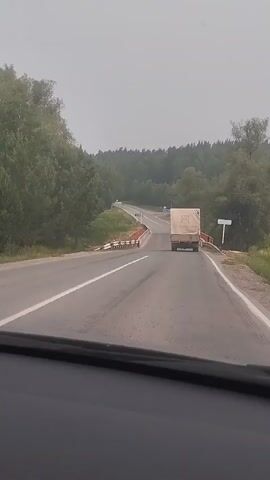 A POV shot from inside a vehicle driving on a two-lane road. The road curves to the left and crosses a bridge. A white truck with a beige tarp is driving on the bridge ahead of the vehicle. There are trees and greenery on either side of the road. The sky is overcast.